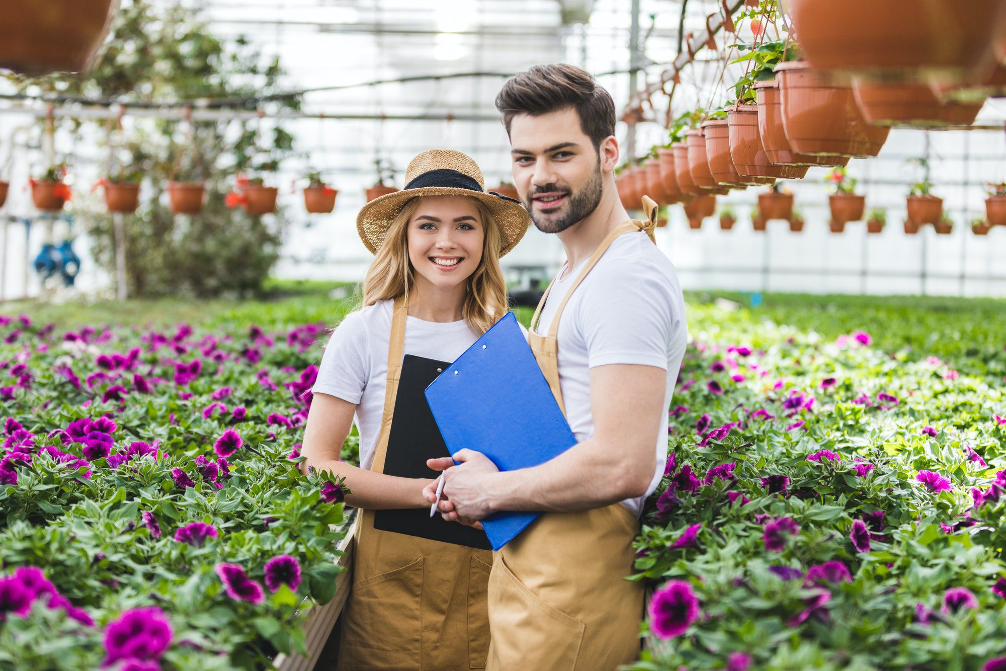 Couple Of Gardeners Holding Clipboards By Flowers In Greenhouse